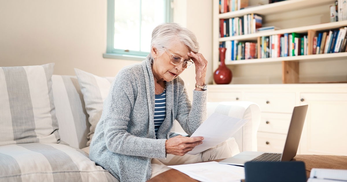 Elderly woman reading a document on a sofa with a laptop nearby.