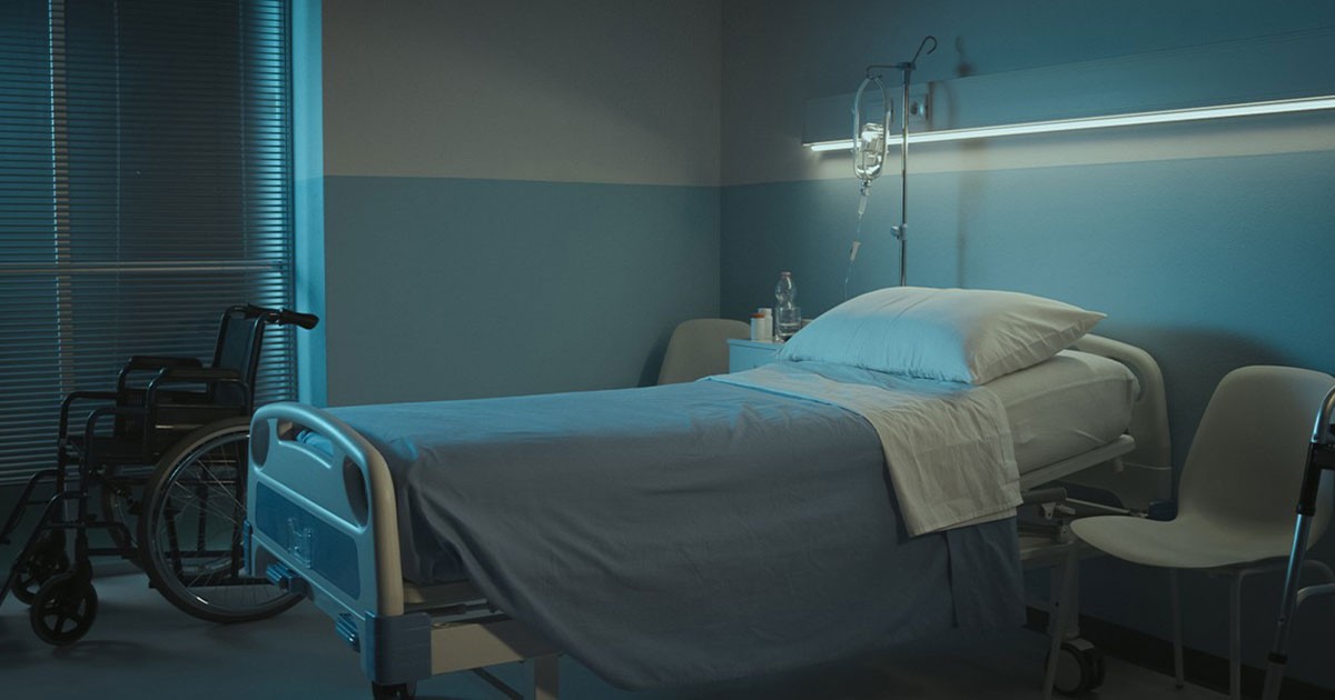 A hospital room with a bed, IV stand, wheelchair, and closed blinds.