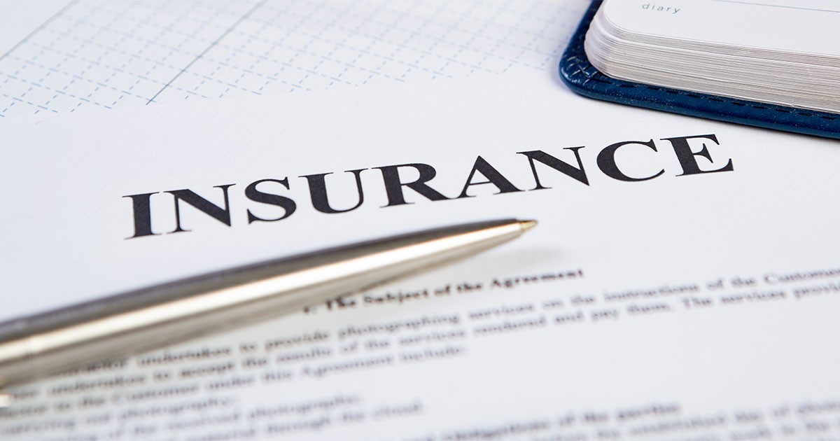 Close-up of an insurance document with the word "INSURANCE" in bold and a pen lying on top. A notebook is partially visible in the background.