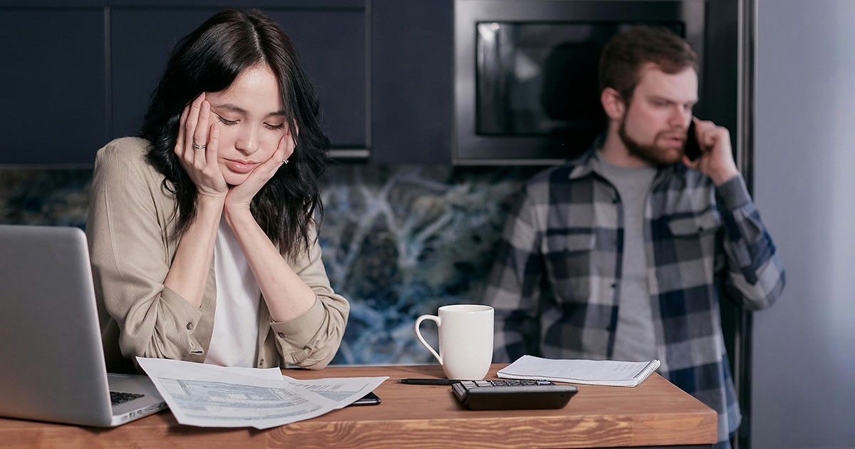 A woman sitting at a table with a laptop and papers, looking tired, while a man stands behind her on the phone in a kitchen. Photo by Mikhail Nilov via Pexels